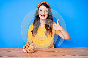 Young beautiful brunette woman sitting on the table eating nachos potato chips smiling happy and positive, thumb up doing