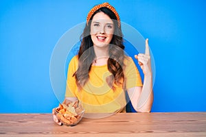 Young beautiful brunette woman sitting on the table eating nachos potato chips surprised with an idea or question pointing finger