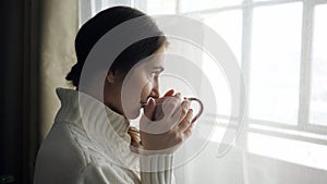 Young beautiful brunette woman in sweater drinking cup of coffee near the window