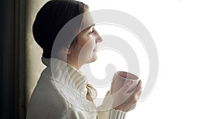 Young beautiful brunette woman in sweater drinking cup of coffee near the window