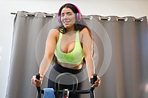 Young and beautiful brunette woman training on the bike in the gym. The girl is listening to music on her headphones. She is