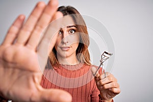 Young beautiful brunette woman using eyelash curler over isolated white background with open hand doing stop sign with serious and