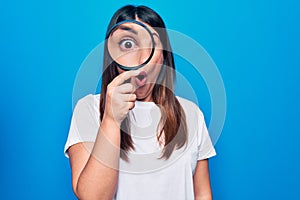 Young beautiful brunette woman using magnifying glass over isolated blue background scared and amazed with open mouth for