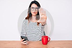 Young beautiful brunette woman using smartphone with open hand doing stop sign with serious and confident expression, defense