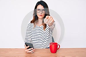 Young beautiful brunette woman using smartphone pointing with finger to the camera and to you, confident gesture looking serious