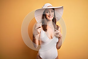 Young beautiful brunette woman on vacation wearing swimsuit and summer hat pointing fingers to camera with happy and funny face