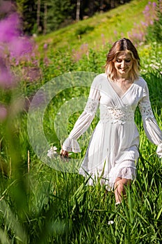 Young beautiful brunette woman walking through wildflowers in alpine meadow, summer dress, mountain, sunlight