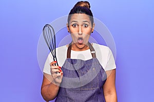 Young beautiful brunette woman wearing apron holding whisk scared and amazed with open mouth for surprise, disbelief face