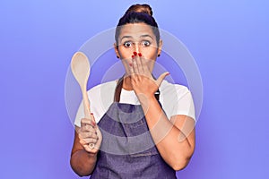 Young beautiful brunette woman wearing apron holding wooden spoon covering mouth with hand, shocked and afraid for mistake