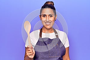 Young beautiful brunette woman wearing apron holding wooden spoon looking positive and happy standing and smiling with a confident