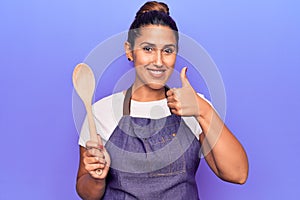 Young beautiful brunette woman wearing apron holding wooden spoon smiling happy and positive, thumb up doing excellent and