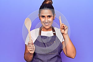 Young beautiful brunette woman wearing apron holding wooden spoon smiling with an idea or question pointing finger with happy