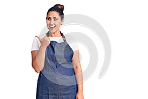 Young beautiful brunette woman wearing apron smiling with happy face looking and pointing to the side with thumb up