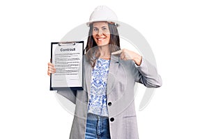 Young beautiful brunette woman wearing architect hardhat holding contract pointing finger to one self smiling happy and proud