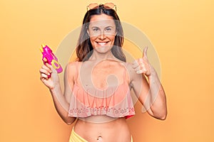 Young beautiful brunette woman wearing bikini playing with water gun over yellow background smiling happy and positive, thumb up