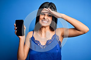 Young beautiful brunette woman wearing blue lingerie holding smartphone showing screen stressed with hand on head, shocked with