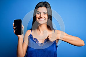 Young beautiful brunette woman wearing blue lingerie holding smartphone showing screen with surprise face pointing finger to