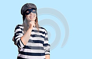 Young beautiful brunette woman wearing burglar mask with hand on chin thinking about question, pensive expression