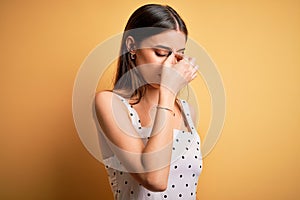 Young beautiful brunette woman wearing casual dress standing over yellow background tired rubbing nose and eyes feeling fatigue