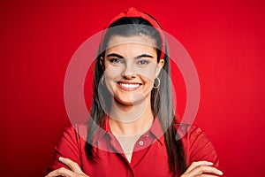 Young beautiful brunette woman wearing casual shirt and diadem over red background happy face smiling with crossed arms looking at