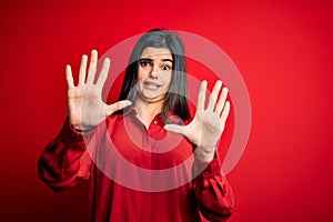 Young beautiful brunette woman wearing casual shirt standing over red background afraid and terrified with fear expression stop