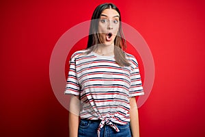 Young beautiful brunette woman wearing casual striped t-shirt standing over red background afraid and shocked with surprise