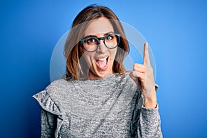 Young beautiful brunette woman wearing casual sweater and glasses over blue background pointing finger up with successful idea