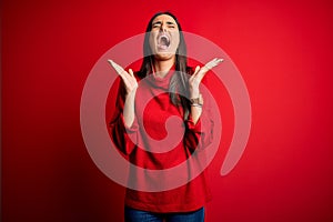 Young beautiful brunette woman wearing casual sweater over isolated red background celebrating mad and crazy for success with arms