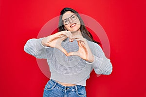 Young beautiful brunette woman wearing casual sweater over red background smiling in love doing heart symbol shape with hands