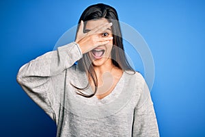 Young beautiful brunette woman wearing casual sweater standing over blue background peeking in shock covering face and eyes with