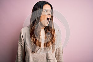 Young beautiful brunette woman wearing casual sweater standing over pink background angry and mad screaming frustrated and