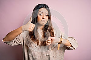 Young beautiful brunette woman wearing casual sweater standing over pink background Doing thumbs up and down, disagreement and