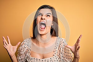 Young beautiful brunette woman wearing casual t-shirt standing over yellow background crazy and mad shouting and yelling with