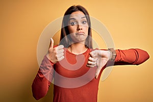 Young beautiful brunette woman wearing casual t-shirt standing over yellow background Doing thumbs up and down, disagreement and