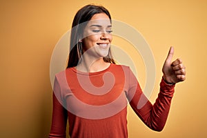 Young beautiful brunette woman wearing casual t-shirt standing over yellow background Looking proud, smiling doing thumbs up