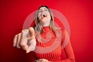 Young beautiful brunette woman wearing casual turtleneck sweater over red background laughing at you, pointing finger to the