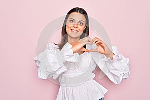 Young beautiful brunette woman wearing casual white dress standing over pink background smiling in love showing heart symbol and