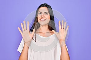 Young beautiful brunette woman wearing casual white t-shirt over purple background showing and pointing up with fingers number ten