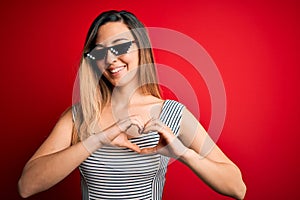 Young beautiful brunette woman wearing funny thug life sunglasses over red background smiling in love showing heart symbol and