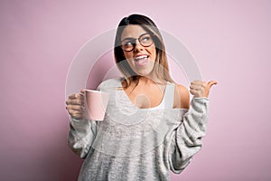 Young beautiful brunette woman wearing glasses drinking pink mug of coffee pointing and showing with thumb up to the side with