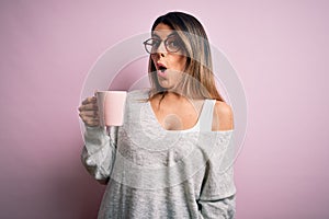Young beautiful brunette woman wearing glasses drinking pink mug of coffee scared in shock with a surprise face, afraid and