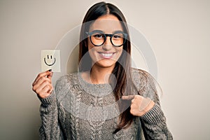 Young beautiful brunette woman wearing glasses holding paper with smile emoji with surprise face pointing finger to himself