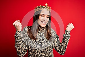 Young beautiful brunette woman wearing golden crown queen over  red background celebrating surprised and amazed for