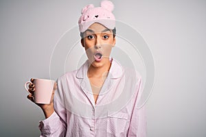 Young beautiful brunette woman wearing pajama and sleep mask drinking cup of coffee scared in shock with a surprise face, afraid
