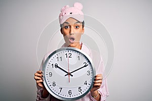 Young beautiful brunette woman wearing pajama and sleep mask holding big clock scared in shock with a surprise face, afraid and