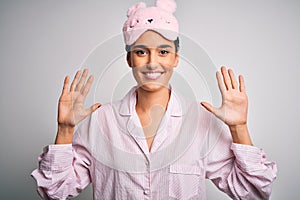 Young beautiful brunette woman wearing pajama and sleep mask over white background showing and pointing up with fingers number ten