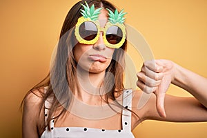 Young beautiful brunette woman wearing pineapple sunglasses over yellow background with angry face, negative sign showing dislike