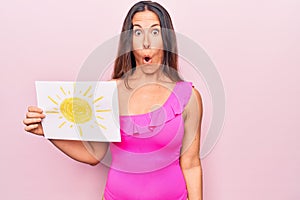 Young beautiful brunette woman wearing pink swimwear holding paper with sun draw scared and amazed with open mouth for surprise,