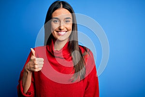 Young beautiful brunette woman wearing red casual sweater over isolated blue background doing happy thumbs up gesture with hand