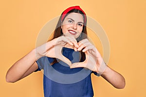 Young beautiful brunette woman wearing red lips over yellow background smiling in love doing heart symbol shape with hands